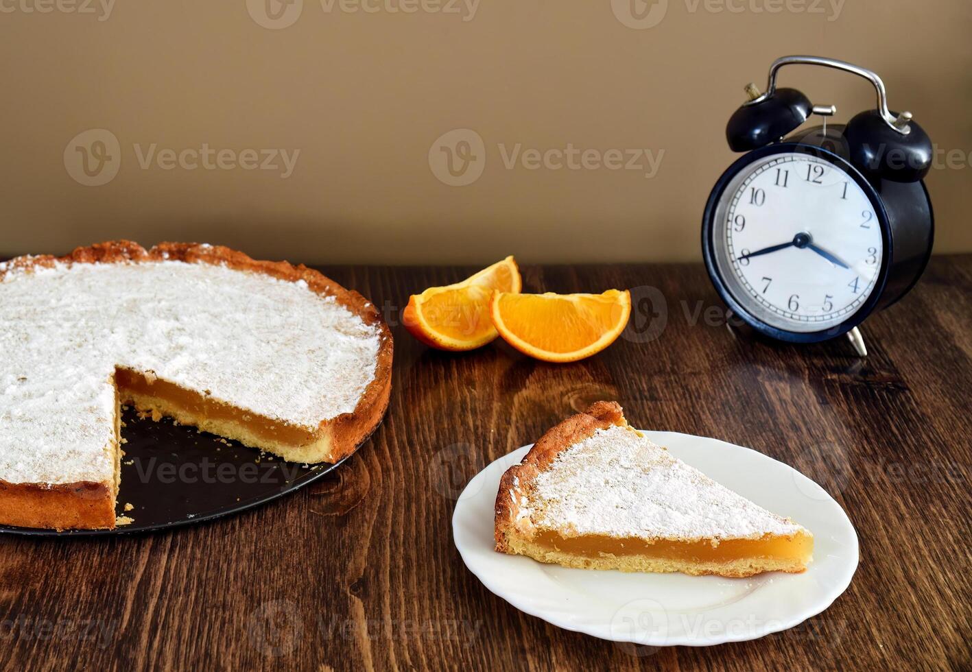 Piece of homemade pie with lemon and orange filling, covered with powdered sugar on a wooden table near orange fruit and alarm clock photo