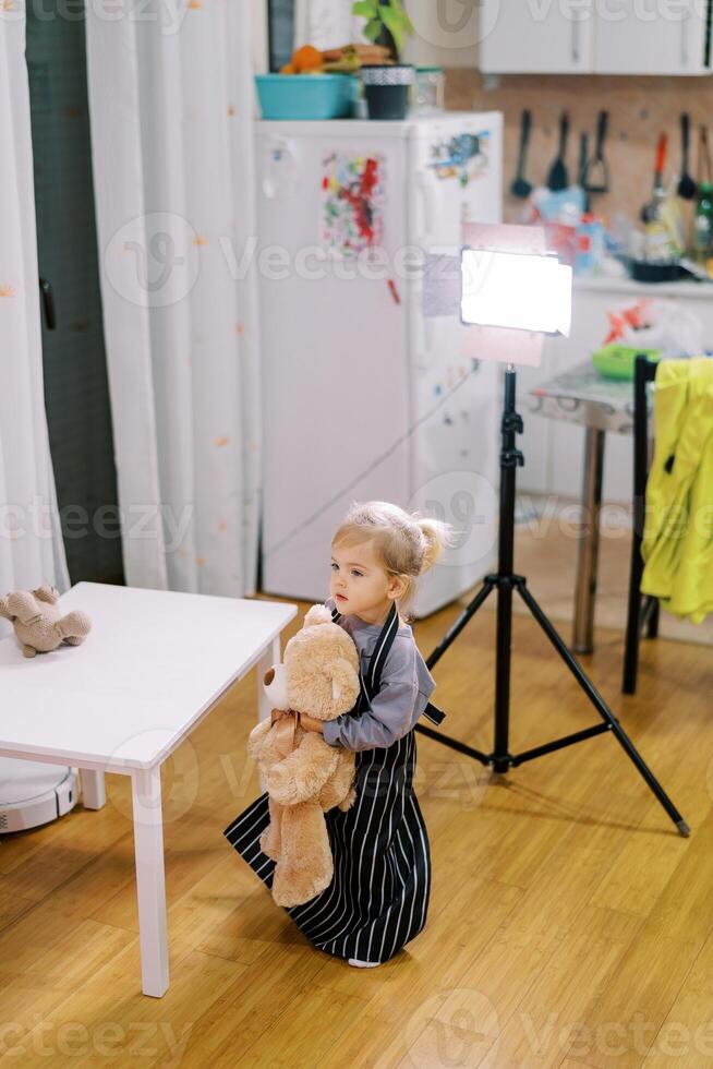 Little girl with a big teddy bear stands near the table in the light of a lamp on a tripod photo