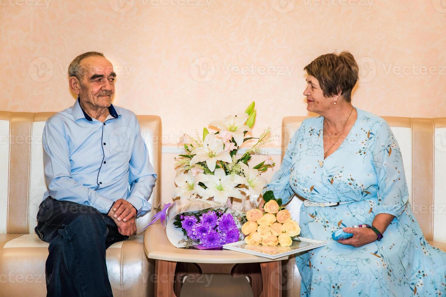 Russian couple over 70, celebrating their golden wedding anniversary at the registry office, sit together on sofas. They smile at each other, surrounded by bouquets of lilies, roses, and chrysanthemum photo