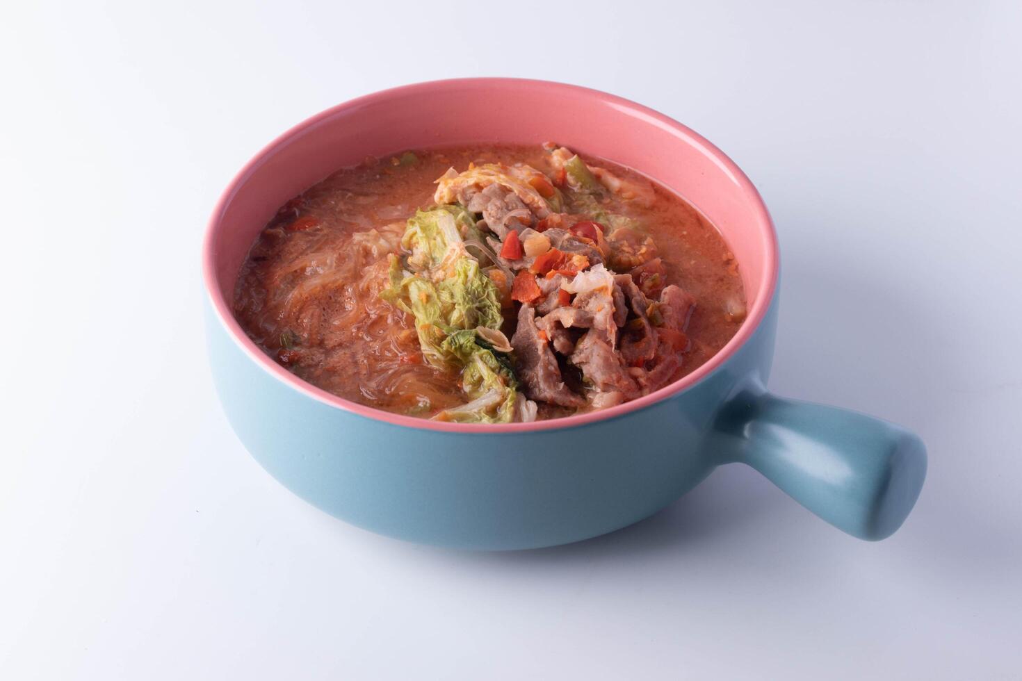 colorful bowl of savory soup with vegetables and meat, featuring pink and blue design, sits on white background. dish includes cabbage, noodles, and rich broth, creating comforting meal photo