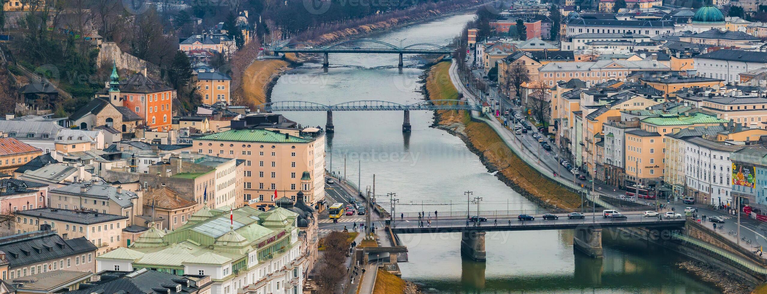 Aerial view of Salzburg, Austria, featuring the Salzach River, multiple bridges, historic pastel buildings, modern structures, and tree lined hills. photo