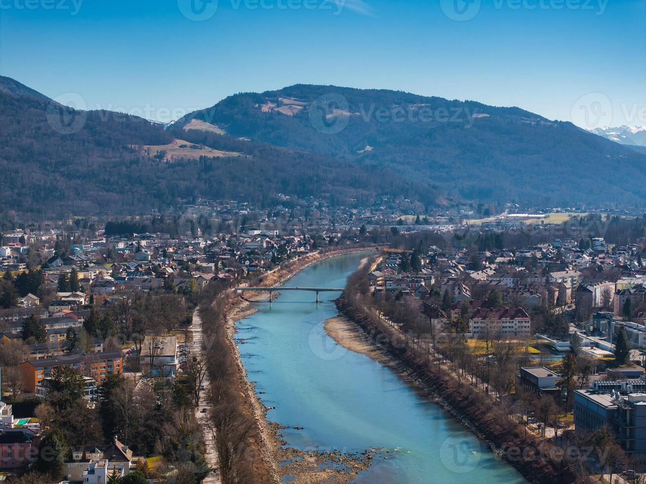 Aerial view of Salzburg, Austria, featuring the Salzach River, a bridge, tree lined paths, urban buildings, and forested mountains under a clear sky. photo