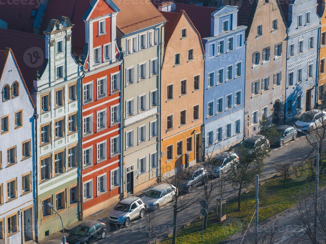 A row of vibrant townhouses in Wroclaw, Poland, with steep roofs, decorative gables, and a tree lined street with parked cars in the foreground. photo