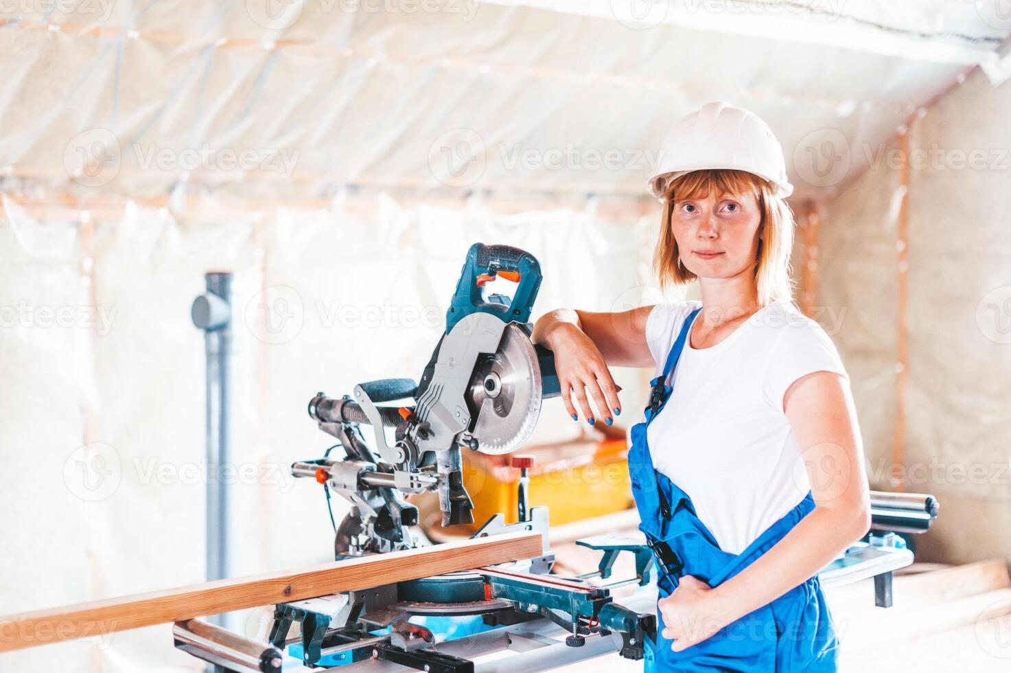 A young woman in a hard hat and overalls stands near a wood cutting machine photo