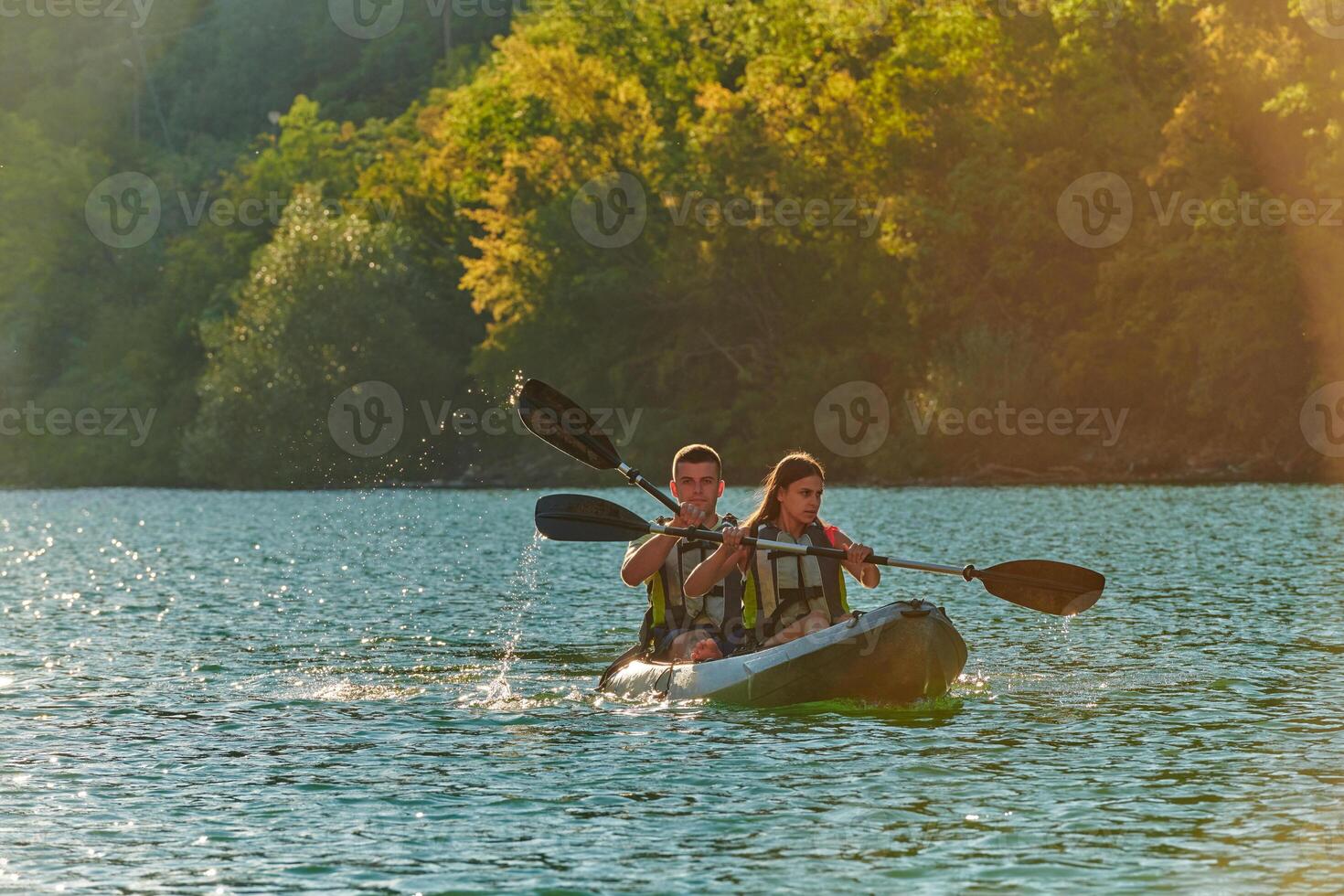 A young couple enjoying an idyllic kayak ride in the middle of a beautiful river surrounded by forest greenery in sunset time photo