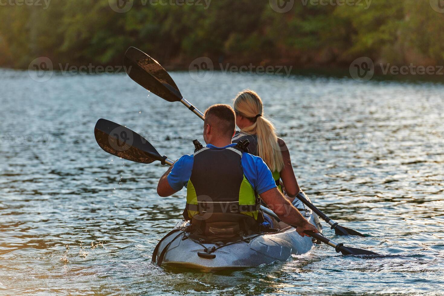 A young couple enjoying an idyllic kayak ride in the middle of a beautiful river surrounded by forest greenery in sunset time photo