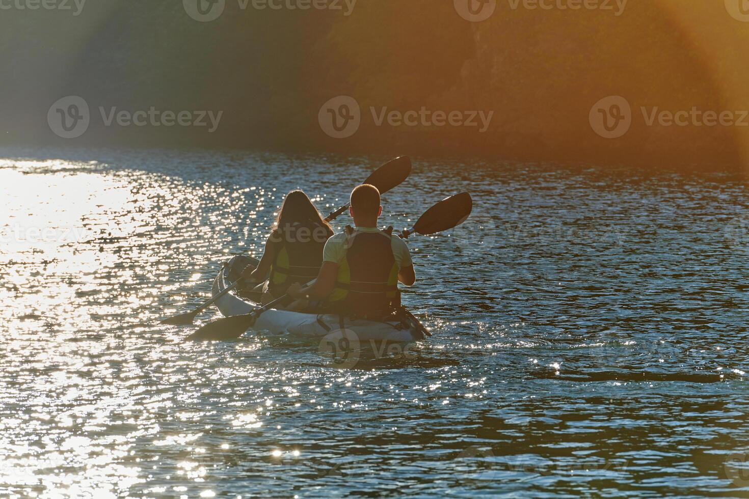 A young couple enjoying an idyllic kayak ride in the middle of a beautiful river surrounded by forest greenery in sunset time photo