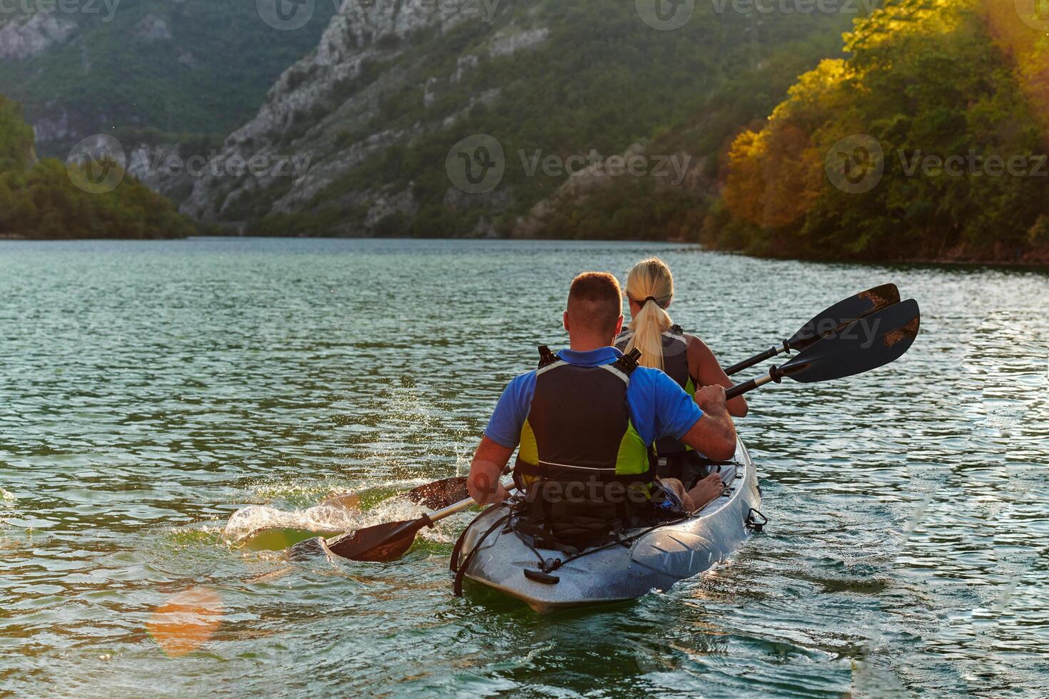A young couple enjoying an idyllic kayak ride in the middle of a beautiful river surrounded by forest greenery in sunset time photo