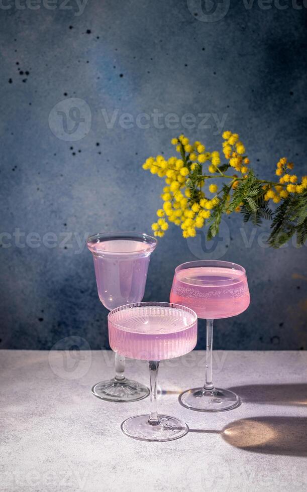Three elegant glasses filled with pink cocktails stand on a table, accompanied by a branch of mimosa flowers, creating a festive atmosphere photo