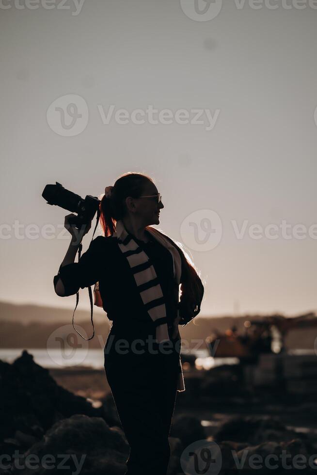 The silhouette of a female photographer confidently holding a camera on her shoulder stands out against the backdrop of a serene sunset. Her long ponytail and stylish scarf add a touch of creativity photo