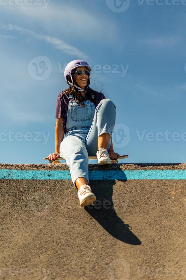 Young latin woman sitting on her surfskate on a pump track ramp wearing helmet and sunglasses photo