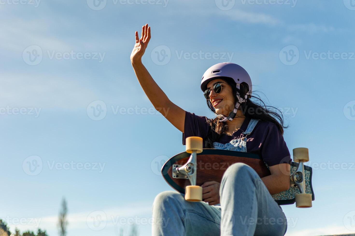 Happy latin surf skater waving while holding her longboard in pump track photo