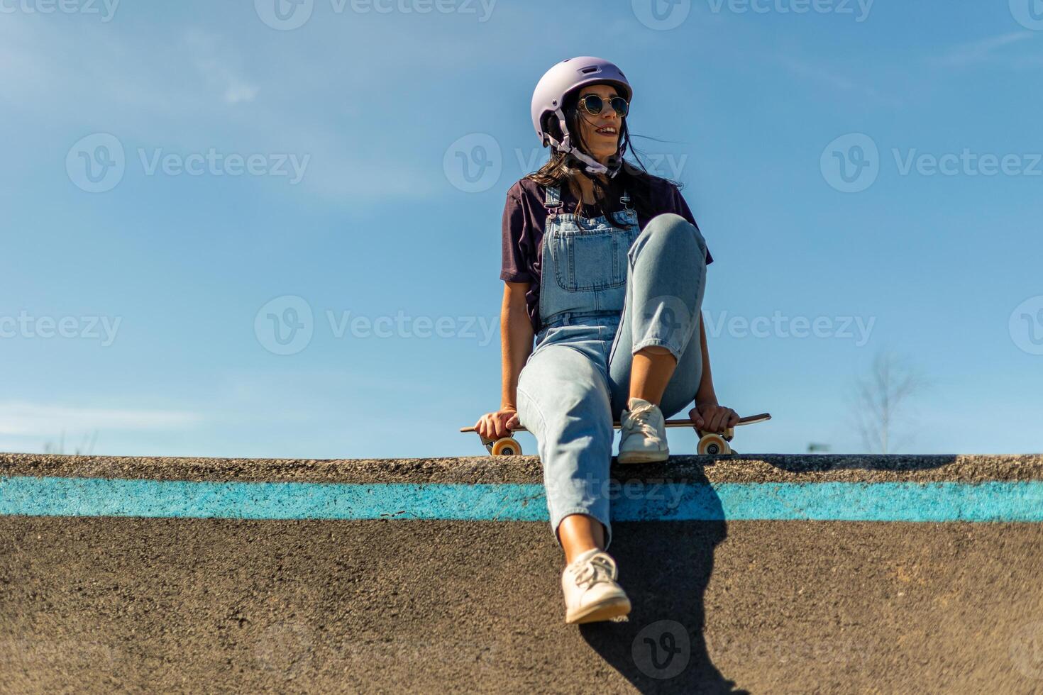Latin skater girl sitting on her surfskate in pump track wearing helmet and sunglasses photo