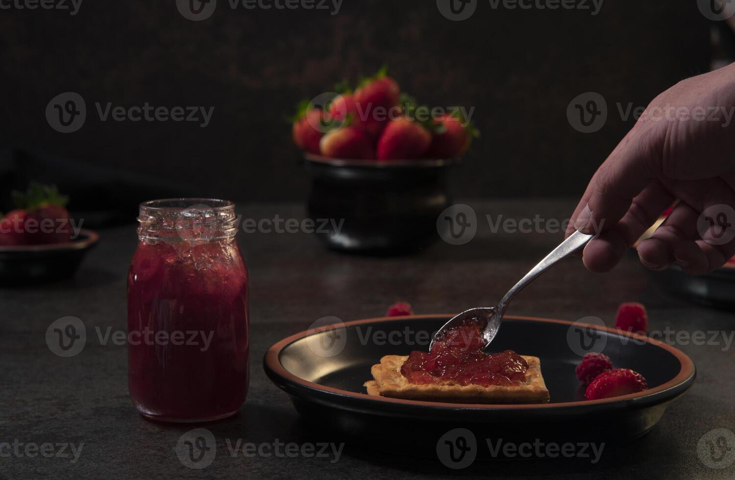 hand with spoon placing strawberry jam on a soda cracker on a black round plate on a black table with a pot of strawberries in the background photo