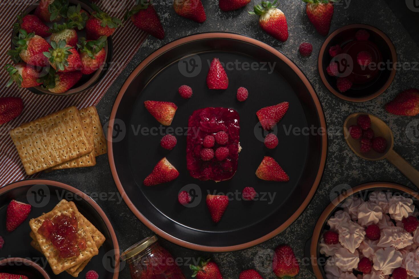 Top view of strawberry and raspberry dessert on a round black ceramic plate surrounded by a pot of strawberries, strawberry jelly, and cookies with strawberry jam photo