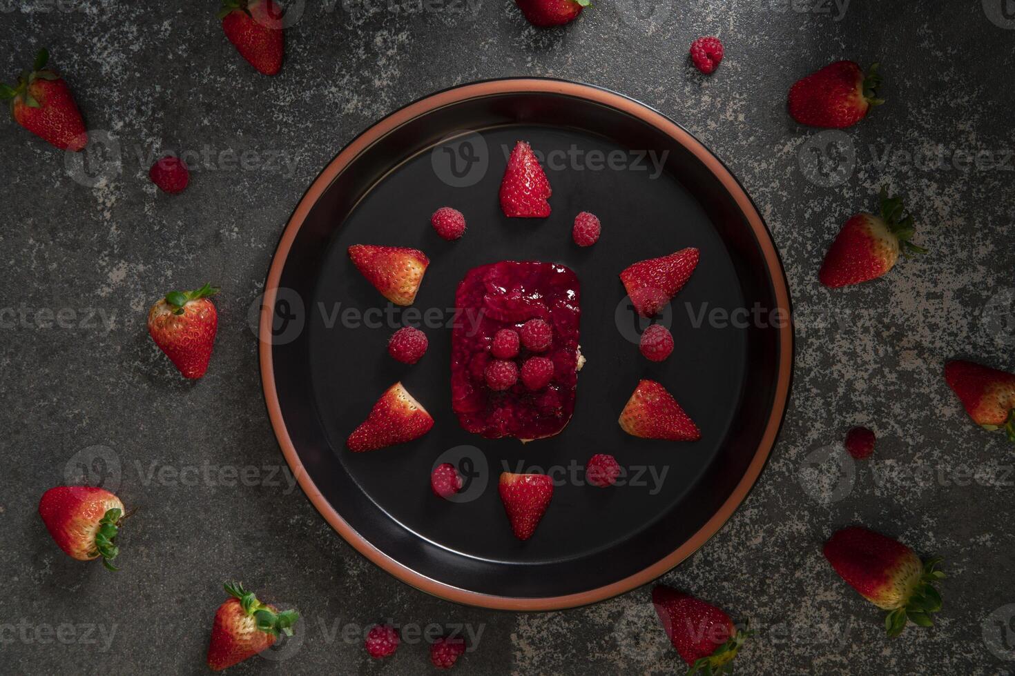 Top view of strawberry and raspberry dessert on a round black ceramic plate surrounded by strawberries photo