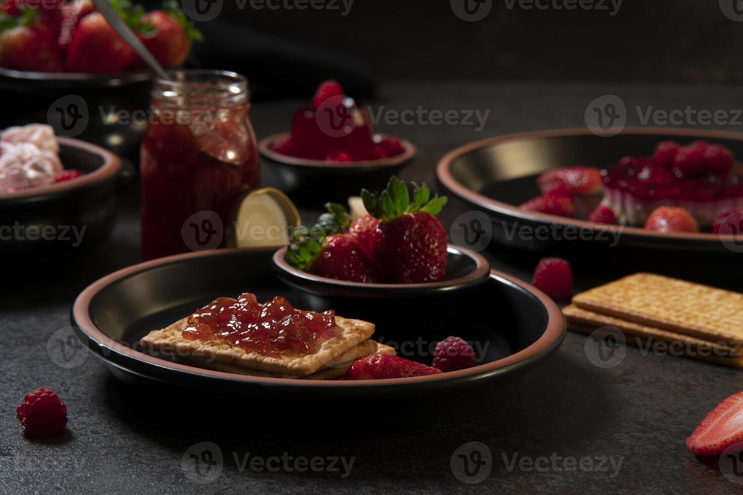 plate with soda crackers with strawberry jam in the foreground against a background of a jam jar and plates with strawberry desserts photo