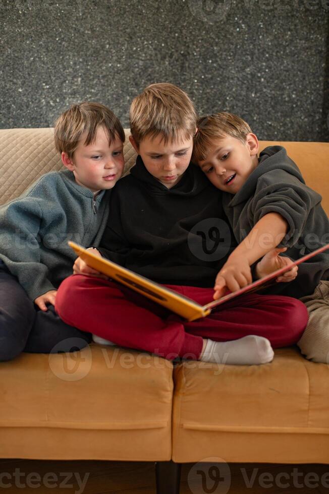 The older brother sits on the sofa, holding a book, while his two younger siblings sit beside him, engaged in the story. photo