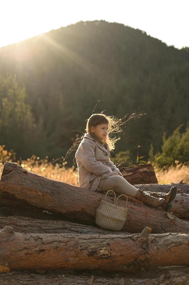 A blonde-haired child enjoys a quiet moment in nature, sitting on wooden logs with sunlight in her hair. photo