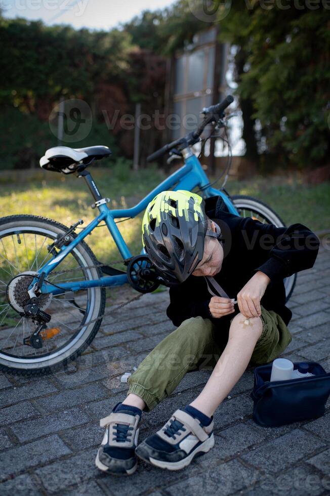 A boy in a bike helmet rests next to his fallen bicycle, using a band-aid to cover a scrape on his knee. photo