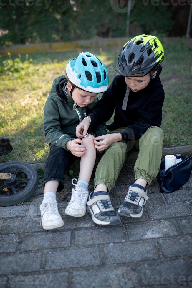 Sitting side by side, two boys in bike helmets focus as the older carefully puts a band aid on the younger ones injured knee photo
