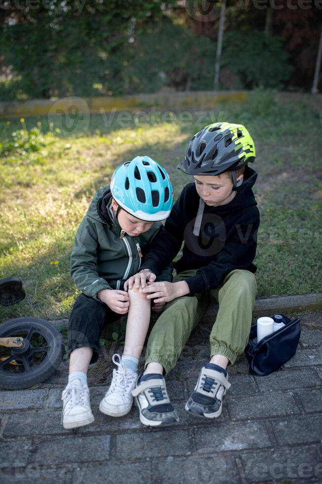 After a small accident, two boys in helmets sit close while the elder helps his younger friend treat a knee scrape. photo