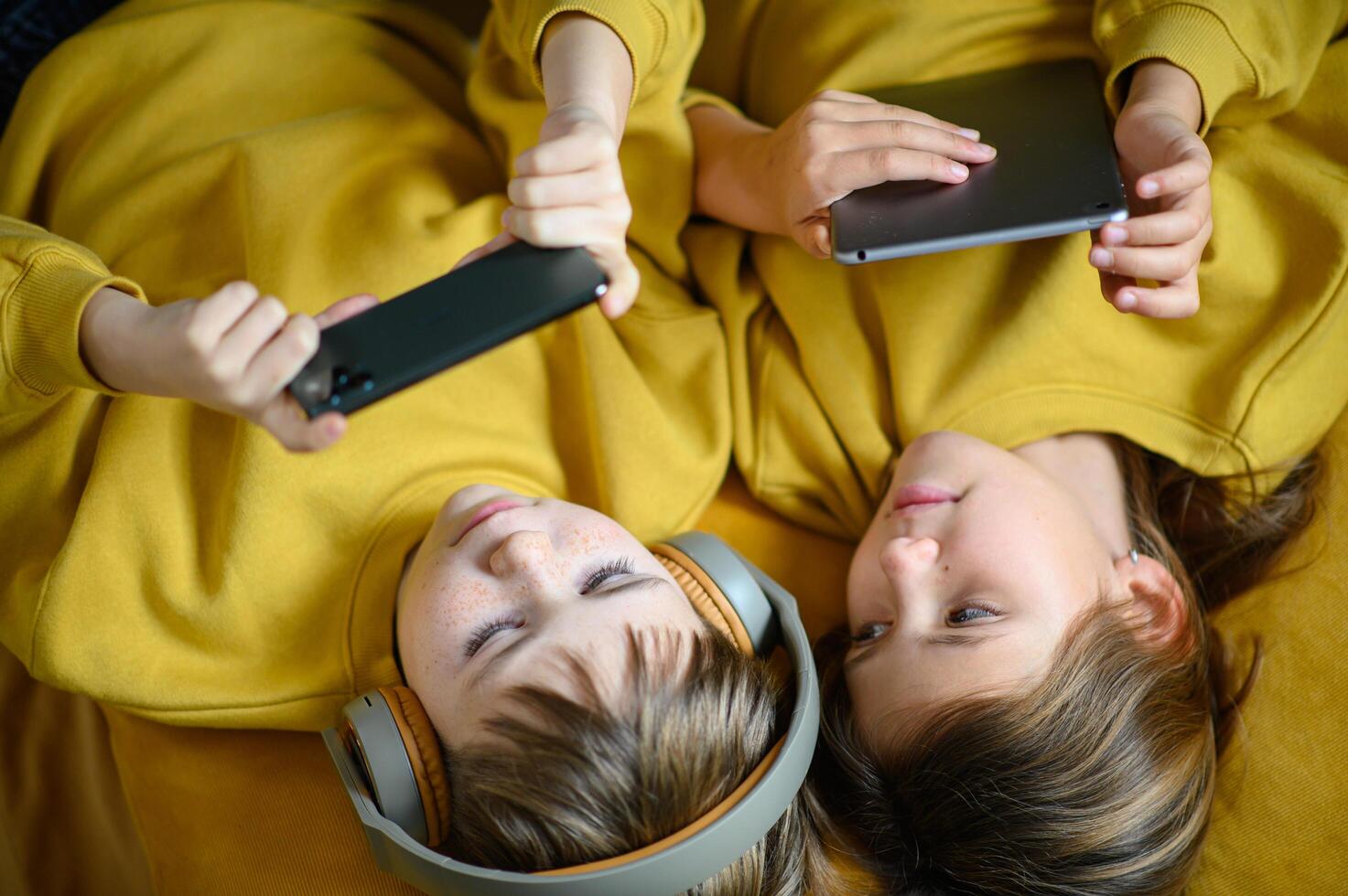 A boy and a girl in yellow lie on the couch, each focused on their smartphone and tablet, wearing headphones. photo