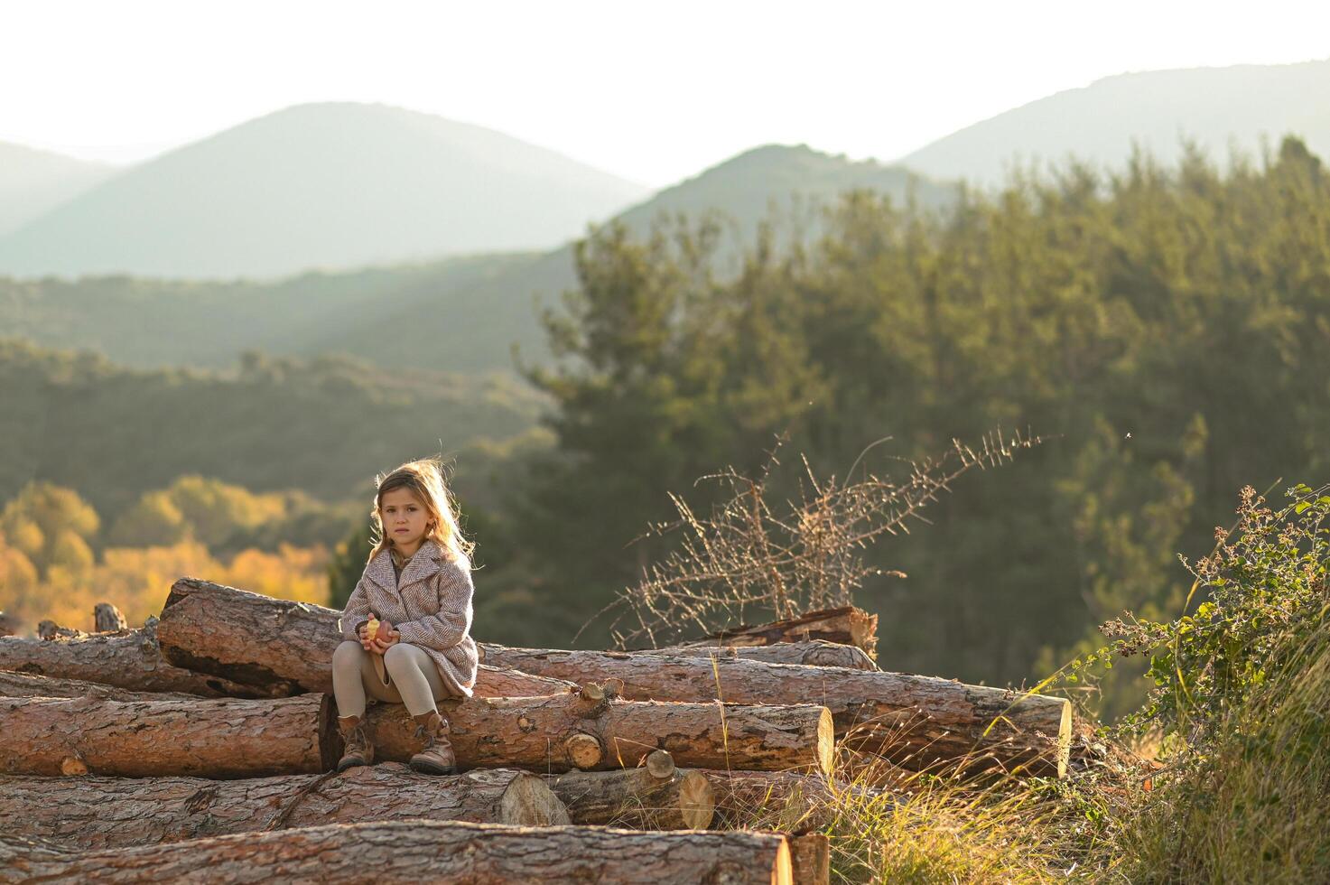 A peaceful moment in nature a little girl sits on rustic wooden logs, holding an apple, surrounded by hills at sunrise photo