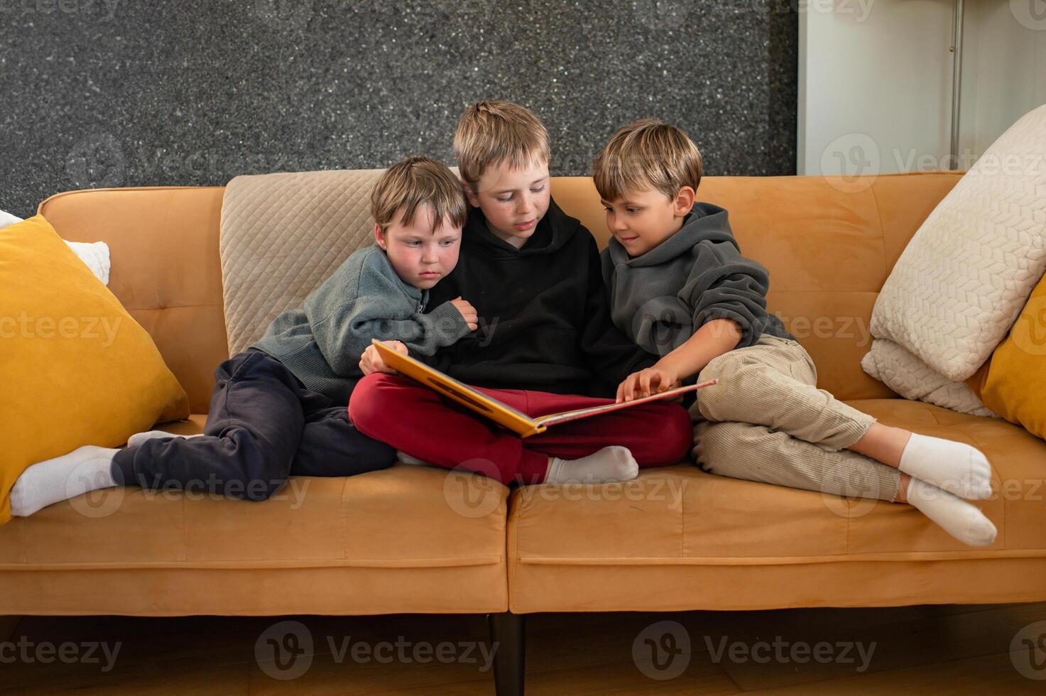 Three boys sit closely on a couch the oldest reads aloud while the younger ones follow along with curiosity. photo