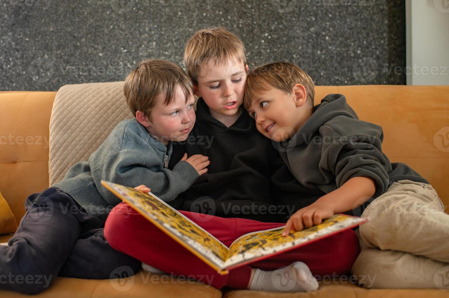 A cozy scene of three boys sitting on a couch, with the eldest reading a book and the younger ones listening intently photo