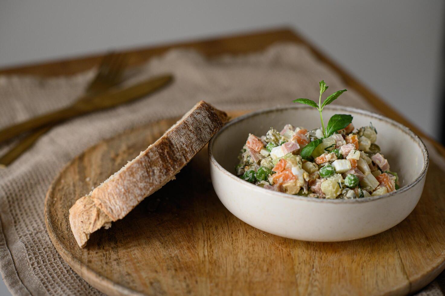 A well-styled food photo featuring Olivier salad, fresh sourdough bread, a fork, and a knife.