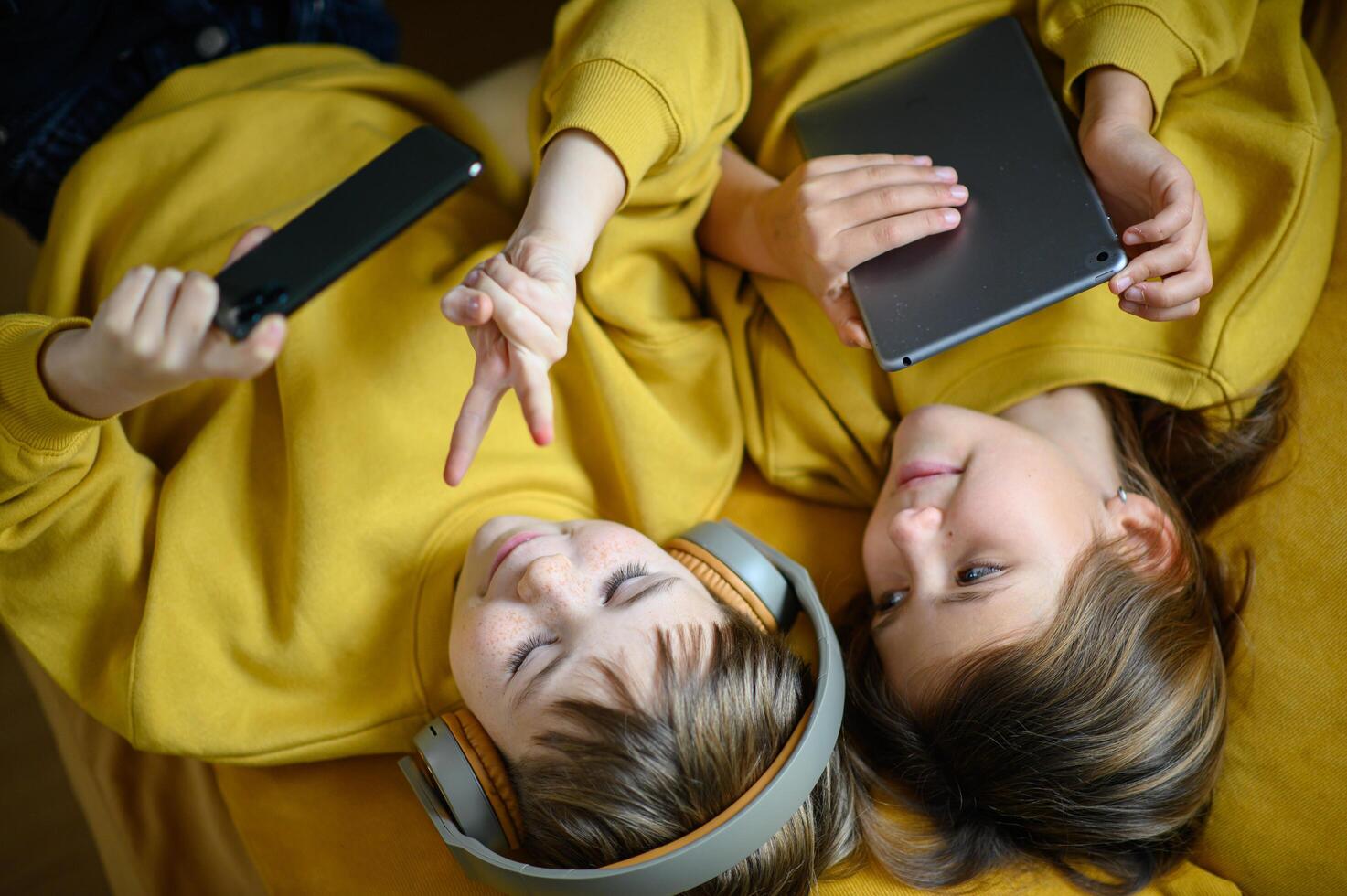 A brother and sister in yellow enjoy their own devices while lying on the couch, lost in music and videos. photo