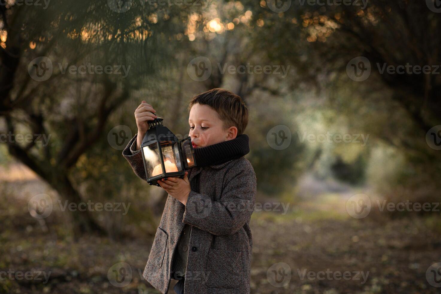 Under the soft glow of the sunset, a boy in a coat blows on his lantern, the olive tree tunnel around him casting a magical atmosphere. photo