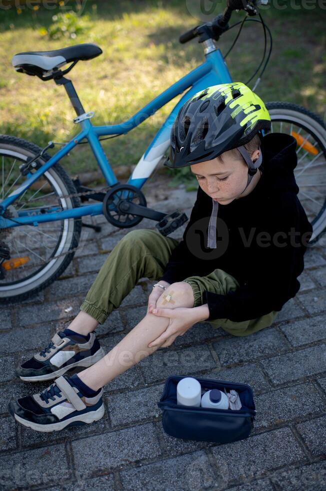 A boy wearing a bike helmet sits on the ground next to his bicycle, carefully applying a band-aid to his scraped knee from a first aid kit he carries with him. photo