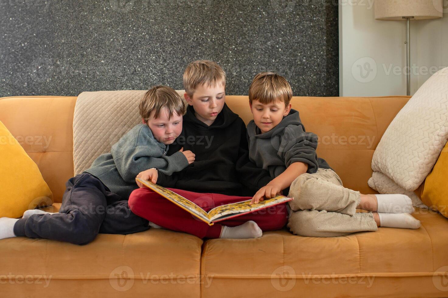A teenage boy sits in the middle of a couch, reading a storybook to his two younger brothers, who lean in eagerly. photo