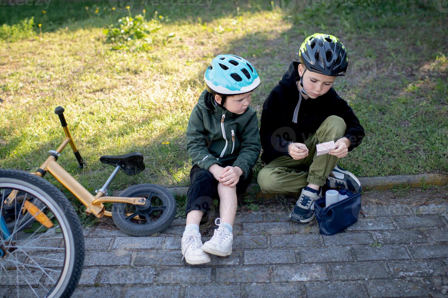 Two boys cyclists wearing helmets are taking a break one is applying a band aid to his scraped knee from a first aid kit photo