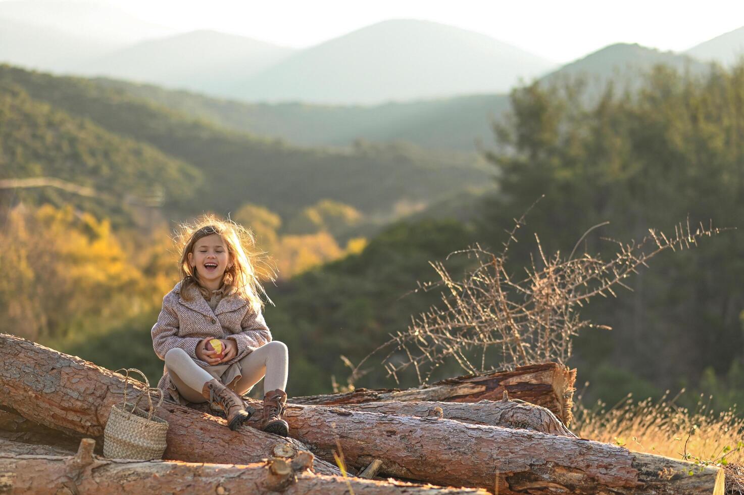 Morning sunlight shines on a young girl eating an apple, seated on wooden logs in a rural, hilly landscape. photo