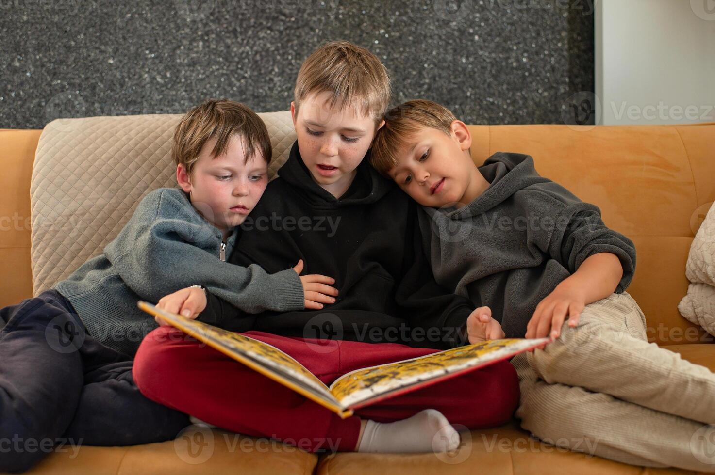 The older brother sits cross-legged on the sofa, reading a fairy tale to his two younger siblings. photo