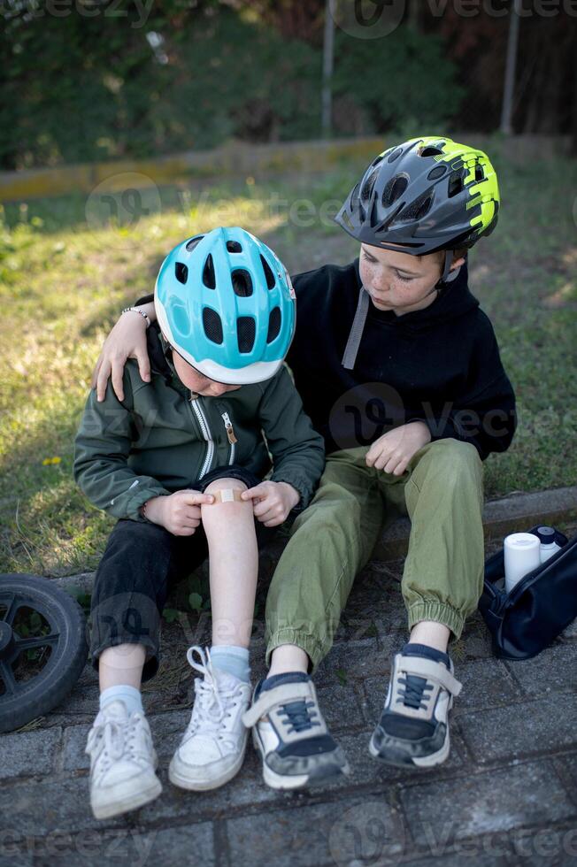 Two brothers or friends in cycling helmets sit together as the older assists the younger in treating his knee injury. photo