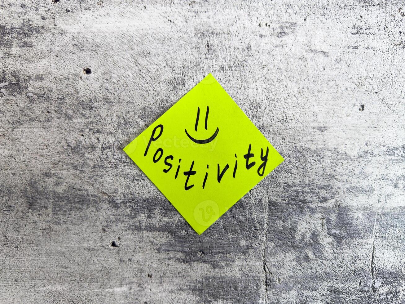 Yellow sticky note in a diamond shape displaying the word Positivity accompanied by a smiley face on a textured grey background, representing the essence of optimistic thinking and cheerful outlook photo