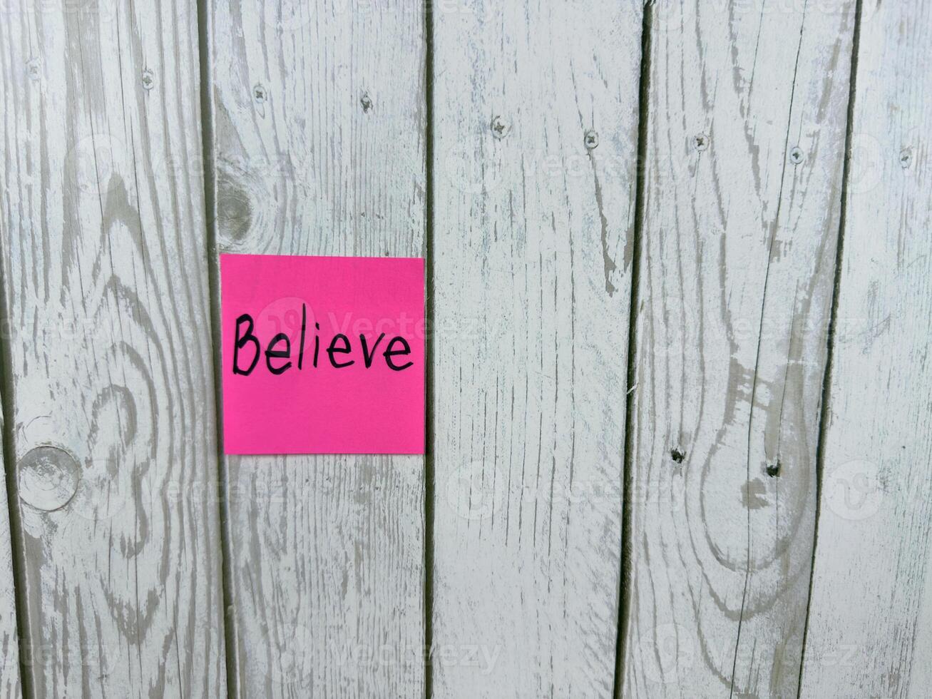 Pink sticky note with the word Believe written on it, pinned on rustic wooden board background. The image represents belief, positivity, and motivation. Ideal for concept themes photo