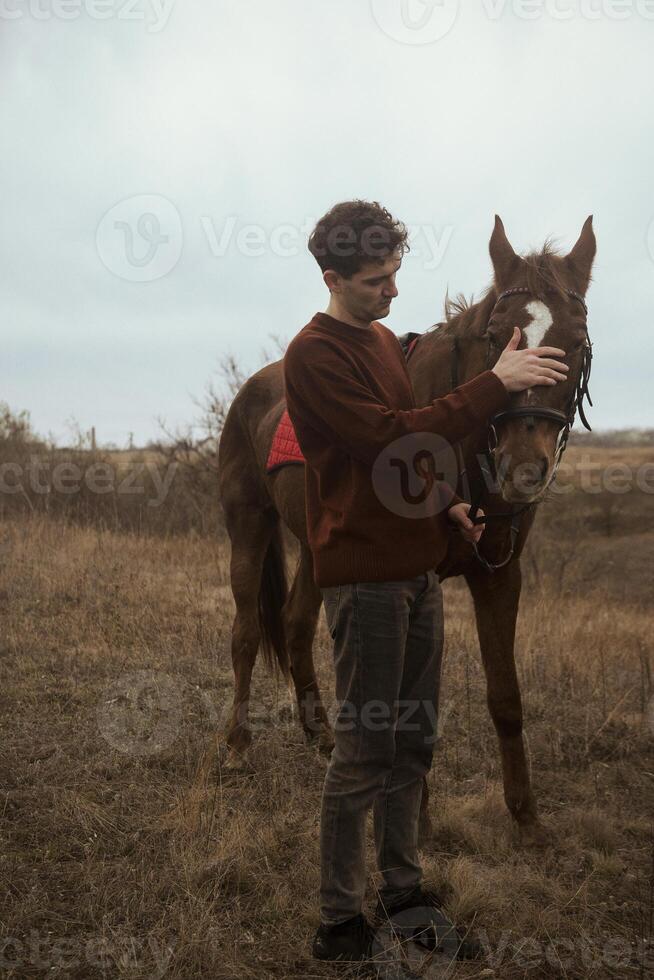 A man in a brown sweater is resting after horse riding with his horse. He is stroking his brown horse. Muchina and horse against the background of nature and a field with autumn grass. Concept of horse riding outdoors, sport and recreation. photo