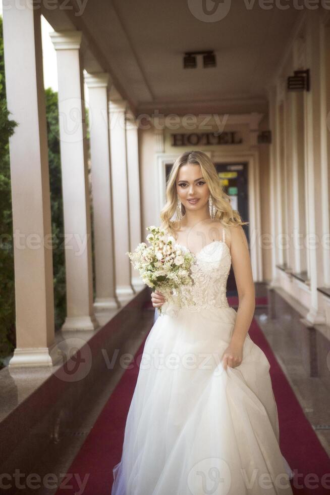 beautiful blonde bride walks along the open gallery of the hotel with a bouquet. long wedding dress, with open shoulders. wavy blonde hair, open shoulders. she smiles and looks at the camera. photo