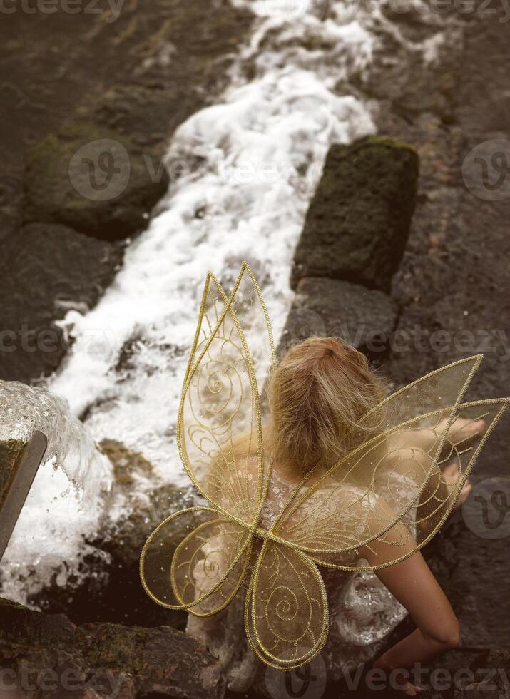 Cute blonde in fairy costume with wings and lace, transparent white dress sits in front of waterfall. Fairy wings are golden, made of transparent chiffon, decorated with embroidery. Top view. photo