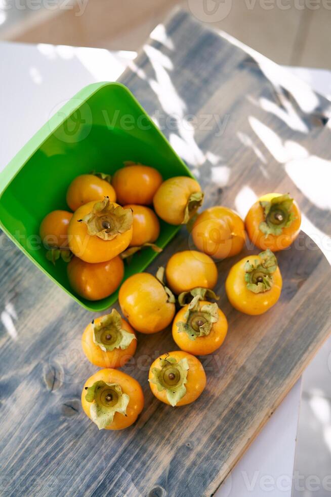 Ripe persimmons spilled out of an overturned square green bowl onto a wooden table. Top view photo