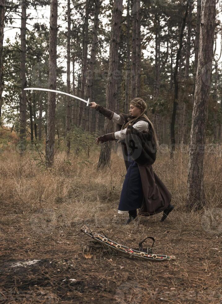 art photo of a warrior girl with a sword in her hands. blonde dressed in a historical costume of a warrior woman, training with a sword in her hands. dressed in a long cloak. warrior raised a sword