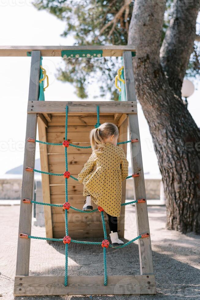 Little girl climbs a rope ladder onto a slide on a playground. Back view photo