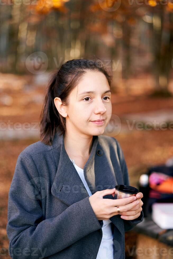 Young woman with a cup of coffee in her hands sits in the autumn forest and looks into the distance photo