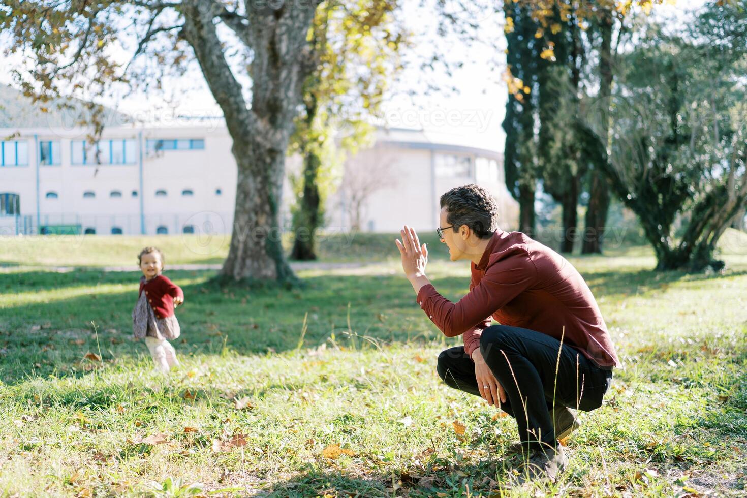 Dad squatting in the park waving his hand to a little girl standing by a tree photo