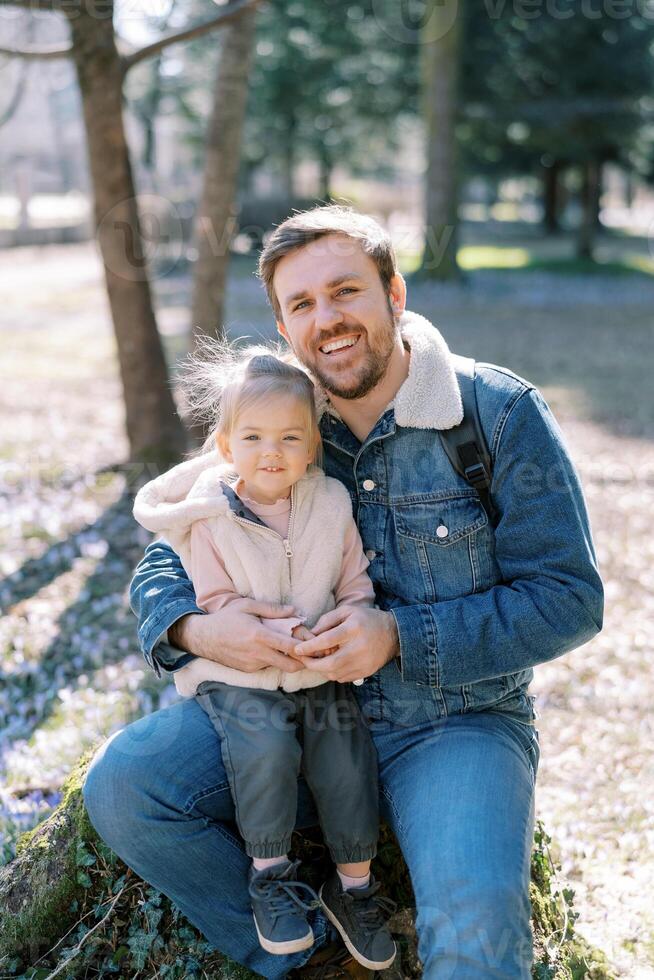 Happy dad with a little girl on his knees sit on a stump in a spring park photo